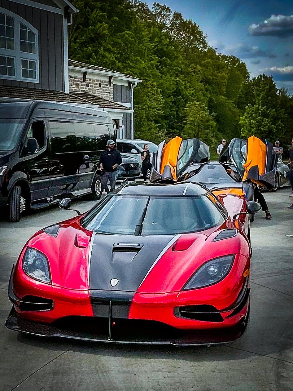 Red and black sports car with gull-wing doors open, parked in a driveway with pe
