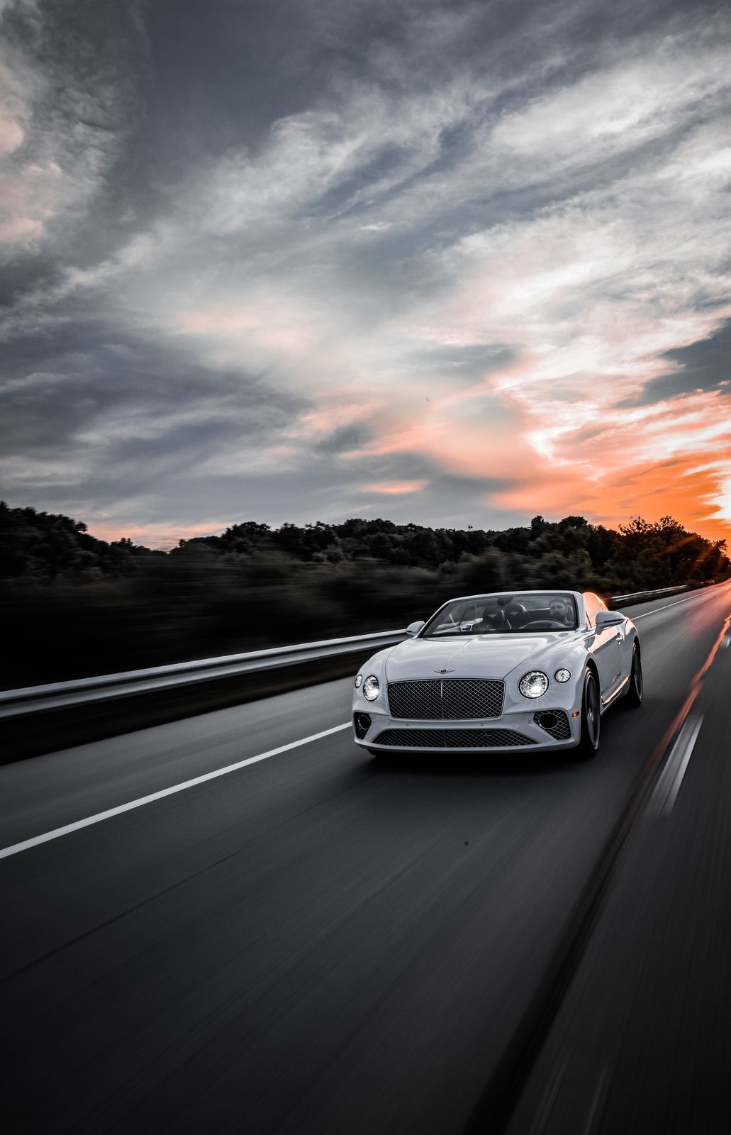 A silver luxury convertible sports car driving on a highway at sunset, with a dr