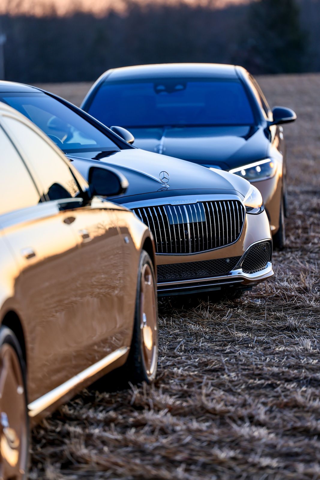 Three luxury cars parked on a dirt field at sunset, including a silver Mercedes-